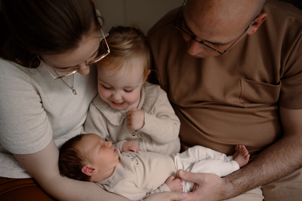 Liefdevol moment tijdens een newbornshoot aan huis: een jong gezin uit Apeldoorn knuffelt hun pasgeboren baby, terwijl de grote zus stralend toekijkt.