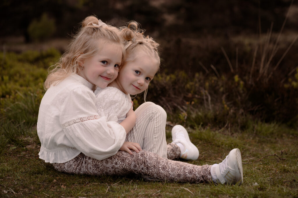 twee zusjes die bij elkaar op de grond zitten en lachend kijken tijdens tijdens een gezinsshoot in de lente