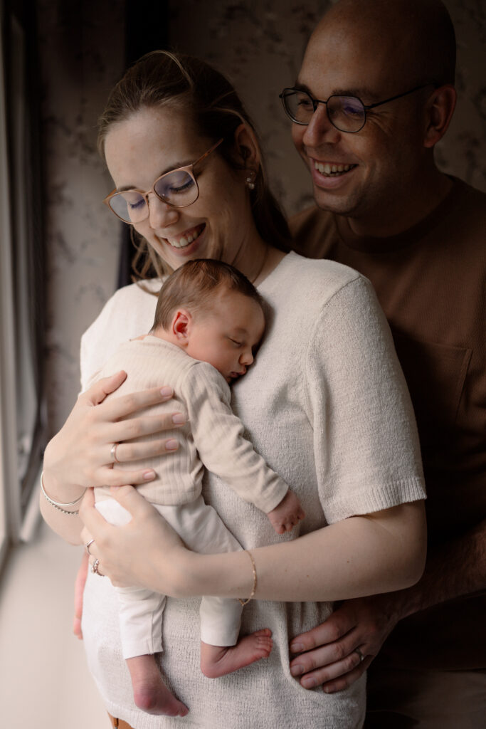 Baby ligt bij moeder op de borst te slapen, terwijl papa lachend over haar schouder kijkt.