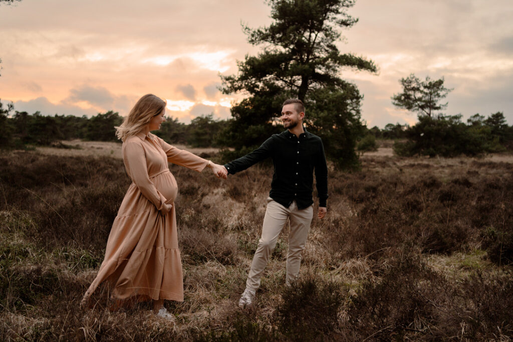 Zwangere dame wordt door haar partner door de heide geleid met een zwangerschapsshoot tijden zonsondergang in de omgeving van Apeldoorn