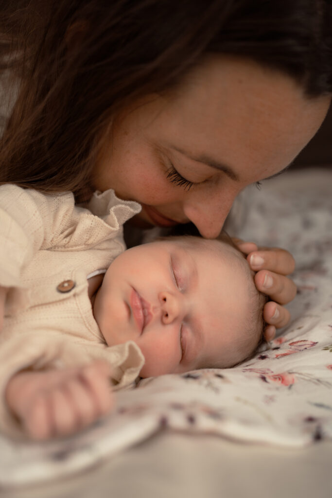 Moeder ligt bij baby op bed en ruikt aan de haartjes van haar kindje tijdens een newbornshoot