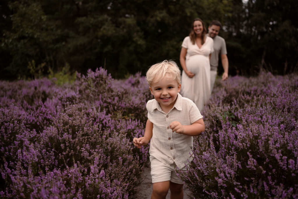 Jongen rent door de bloeiende heide met zijn ouders erachter tijdens een gezinsshoot door Joenne Bokhorst Fotografie